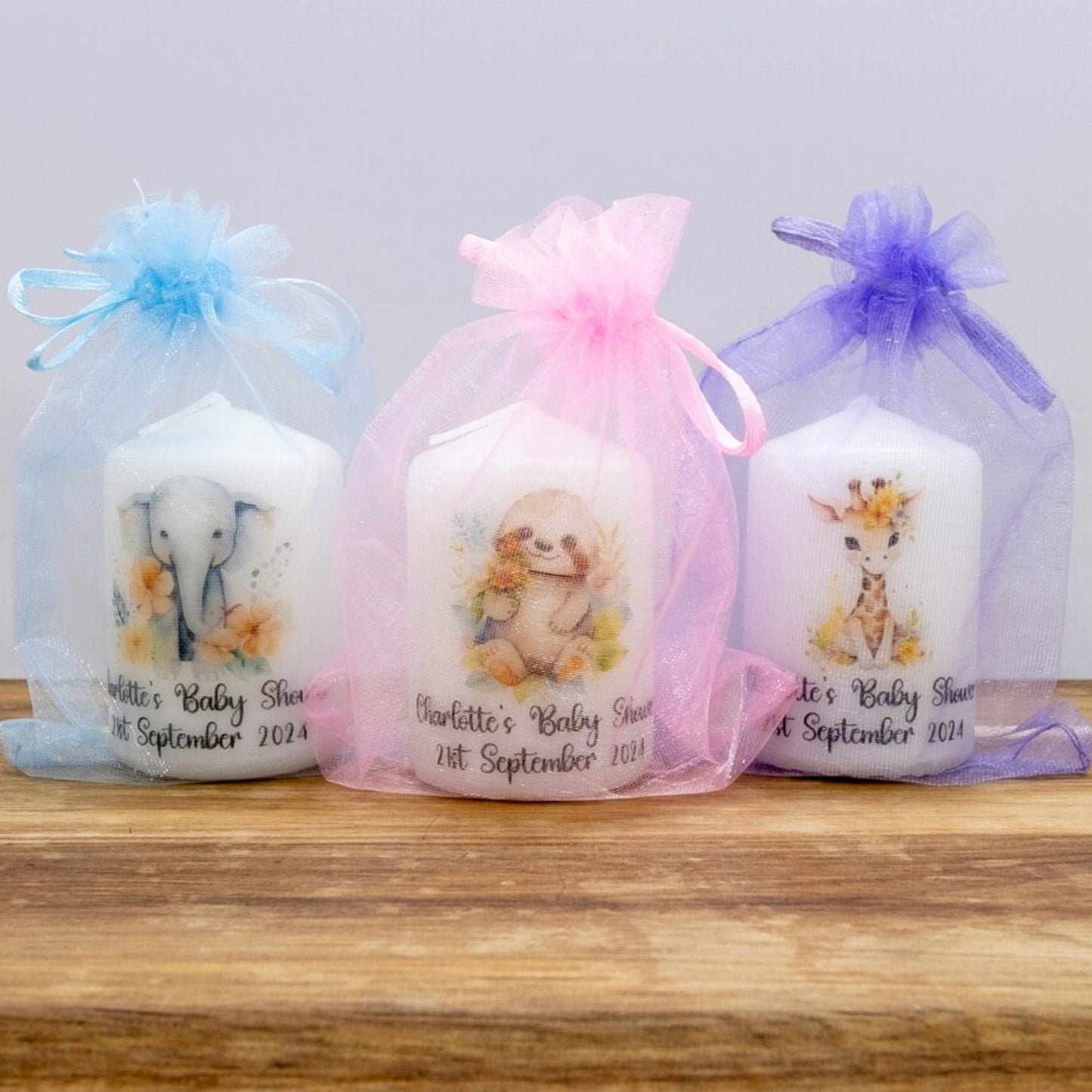 Three baby shower candles in decorative bags on a wooden surface with a light gray background.