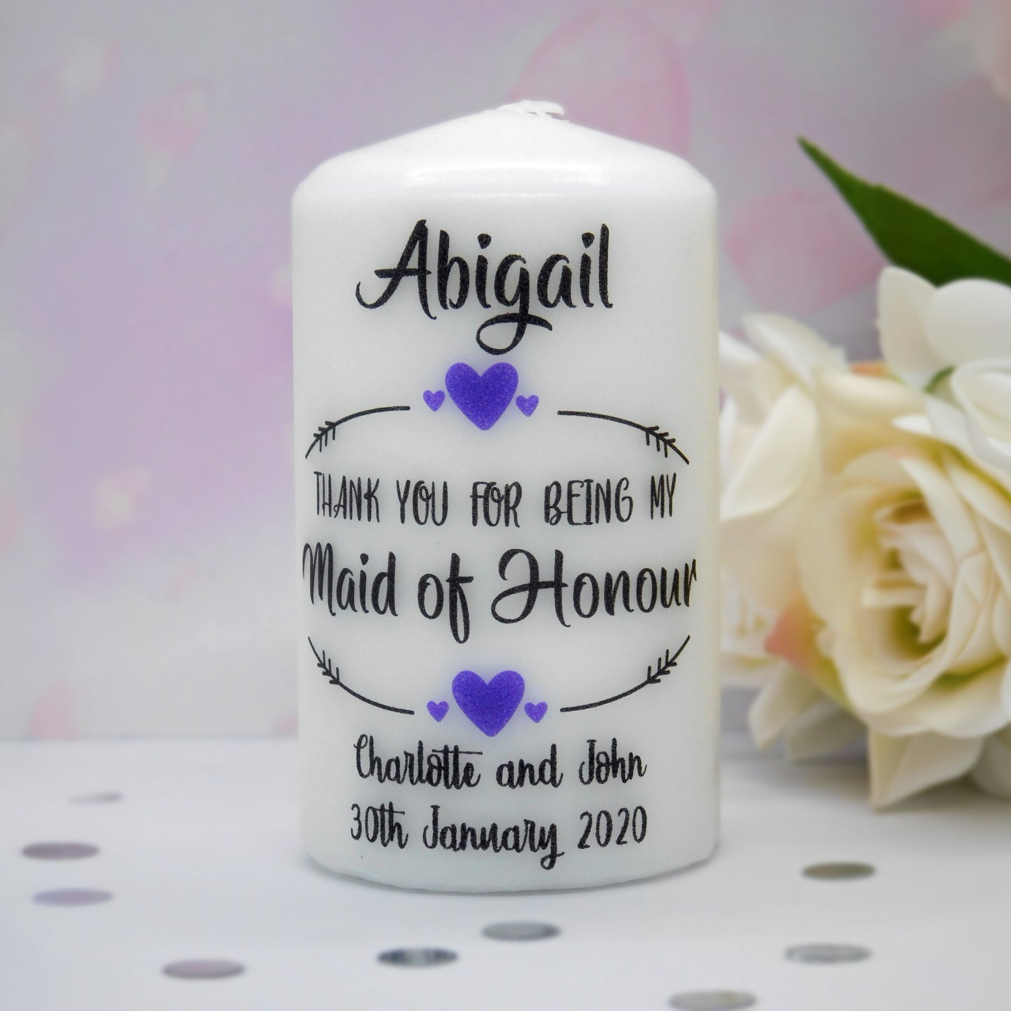 A white pillar candle with personalised black text and purple heart accents, reading 'Abigail Thank You for Being My Maid of Honour Charlotte and John 30th January 2020'. The candle is photographed on a white surface with blurred cream flowers in the background.
