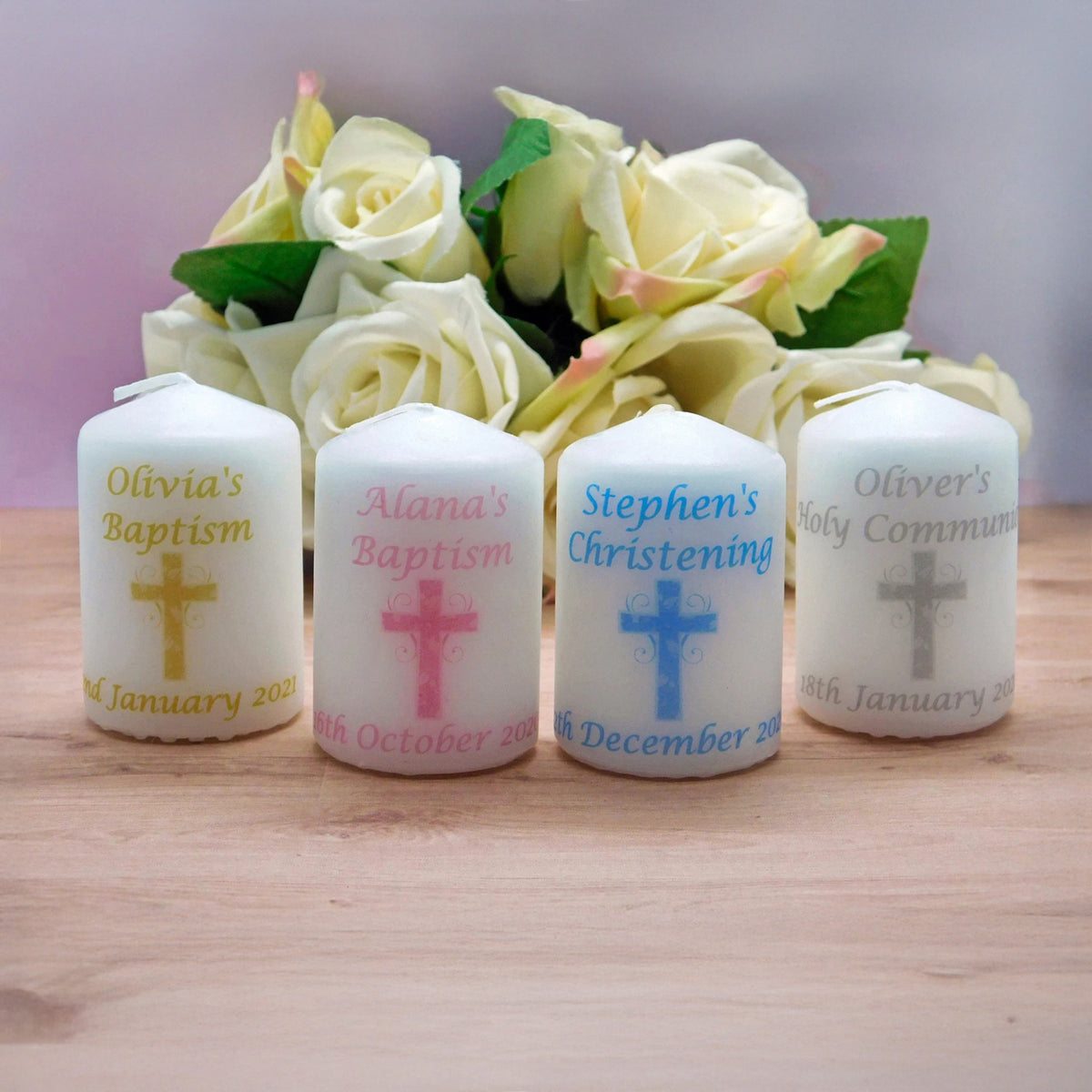 Four white custom-printed pillar candles with Christian symbols and names, arranged in front of a bouquet of cream roses on a wooden surface.