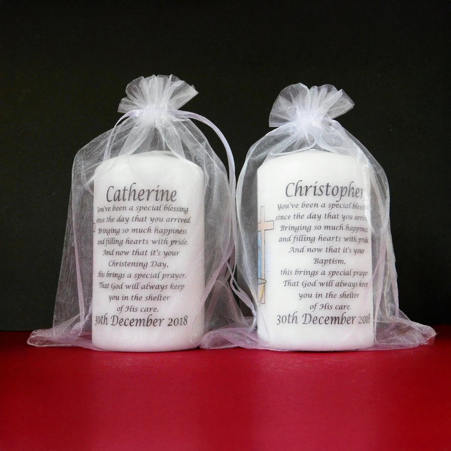 Two white candles in sheer bags with personalized text on a red surface and black background