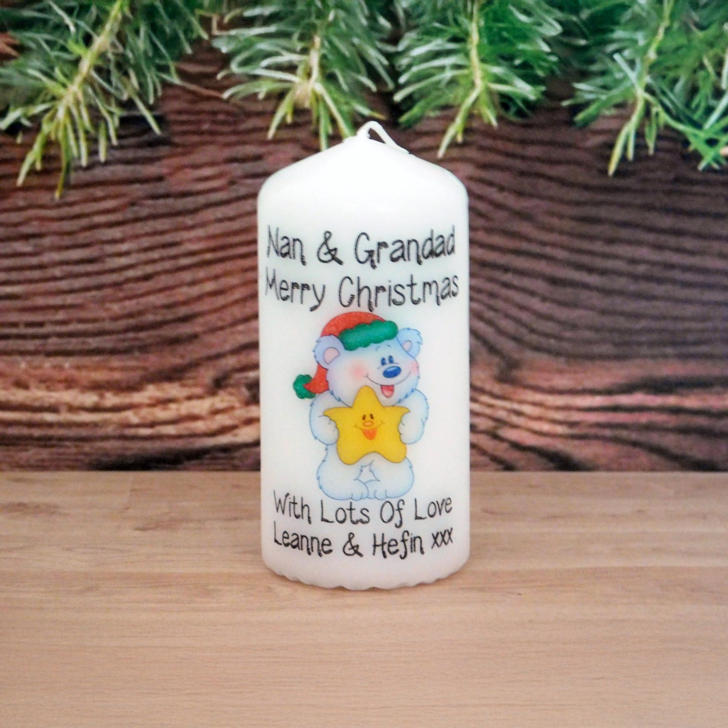 Decorative candle with a cartoon bear design and Christmas message, placed on a wooden surface with a wicker basket and greenery in the background.
