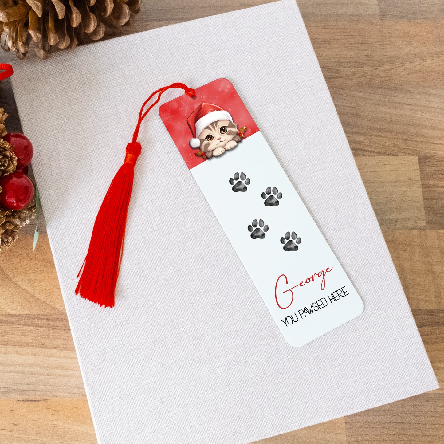 Bookmark with a cat in a Santa hat and paw prints on a white surface with Christmas decorations.