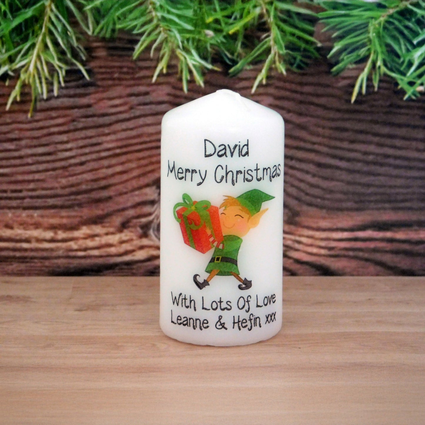 White candle with Christmas design and text on a wooden surface with greenery in the background
