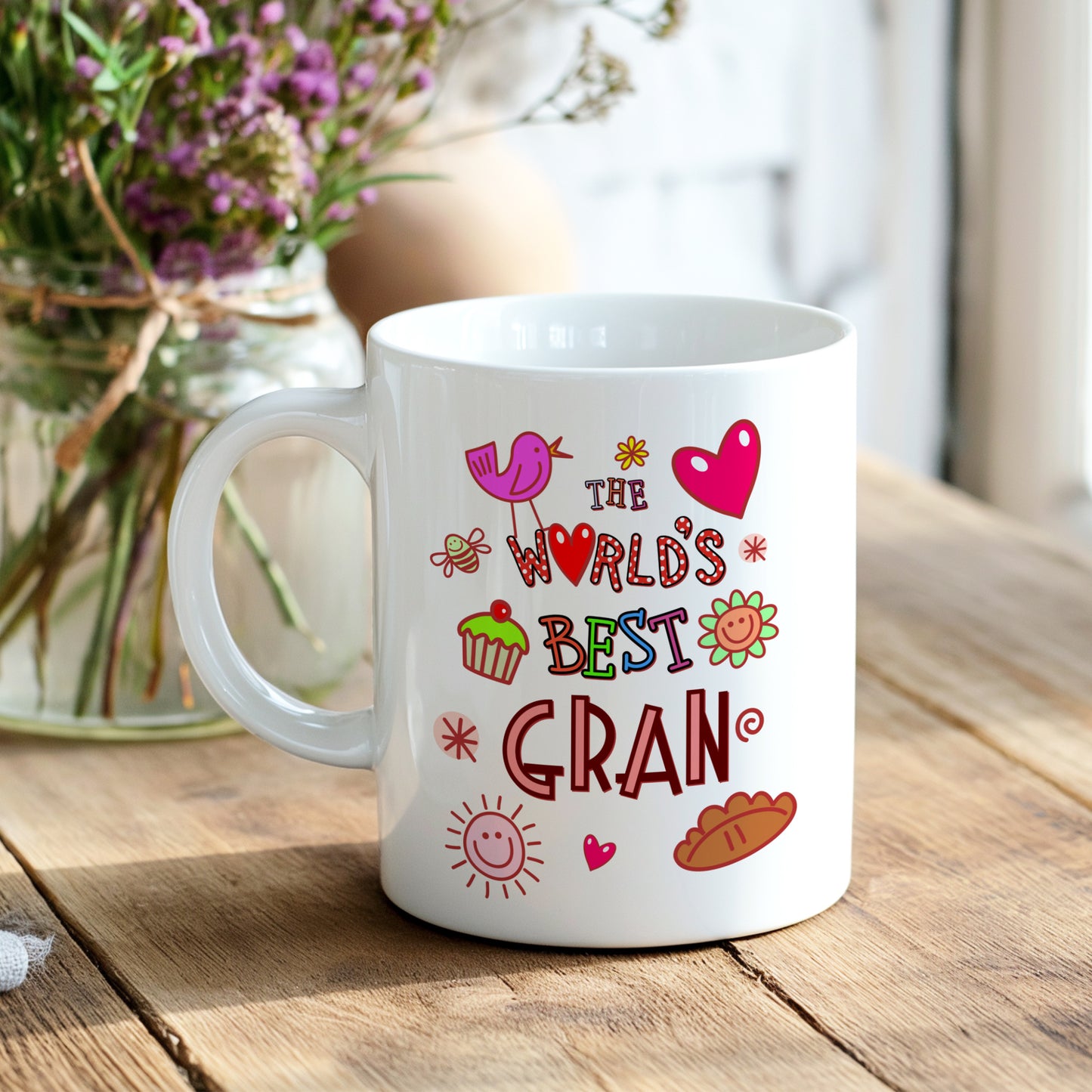 White mug with colorful text 'The World's Best Gran' on a wooden table with flowers in the background