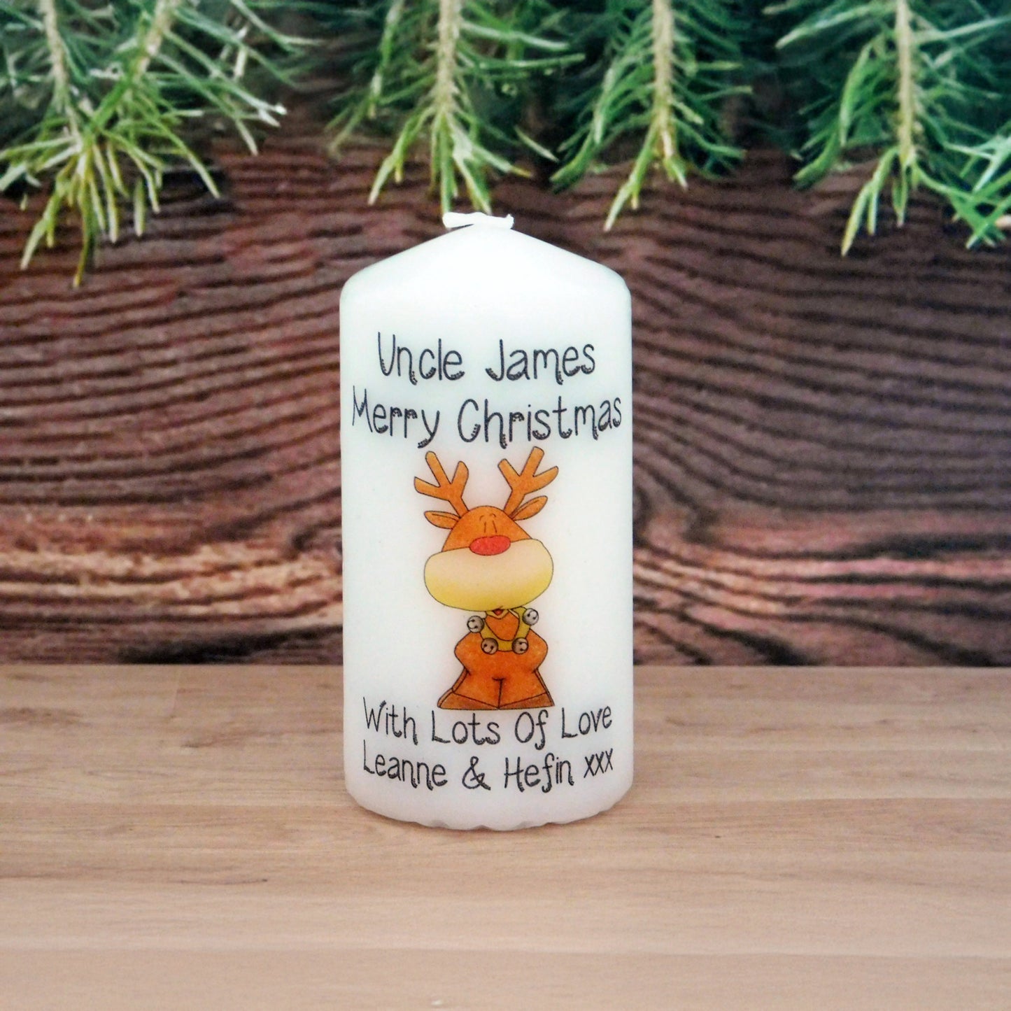 Personalized Christmas candle with a reindeer design and text, placed on a wooden surface with a wicker basket and greenery in the background.