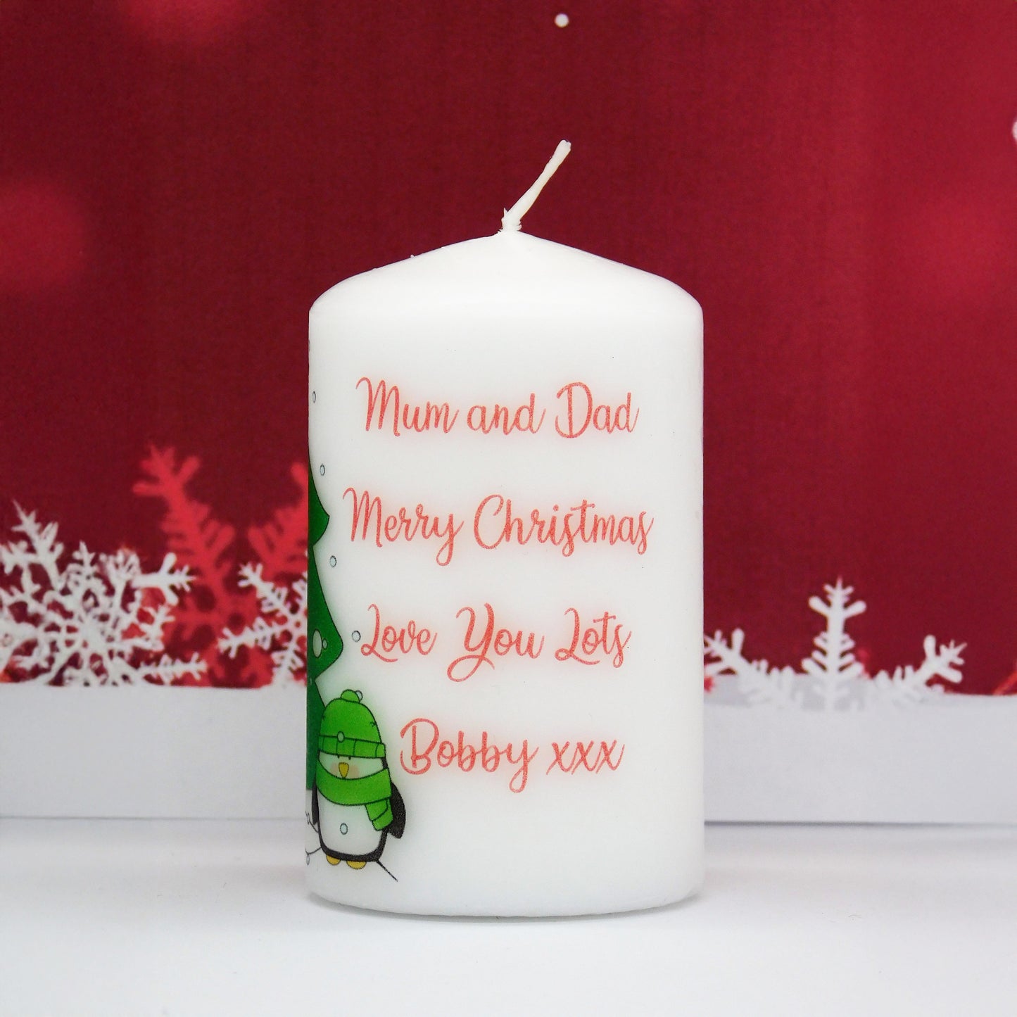 White candle with red text and a penguin design on a red background with snowflakes.