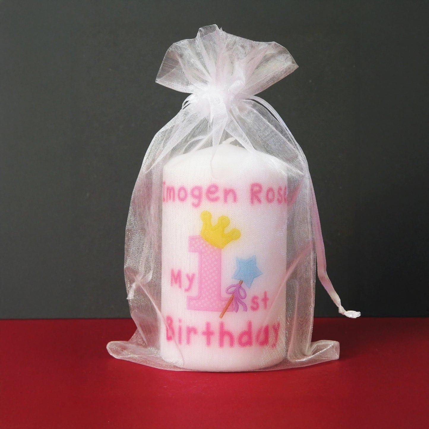 A white candle wrapped in a sheer white organza bag, printed with pink text reading 'Imogen Rose My 1st Birthday' and decorated with a pink number one, yellow crown, and blue star wand, set against a dark grey background and red surface.