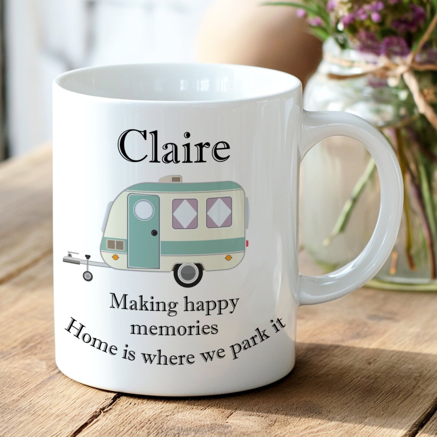 White mug with a camper design and text on a wooden surface with flowers in the background
