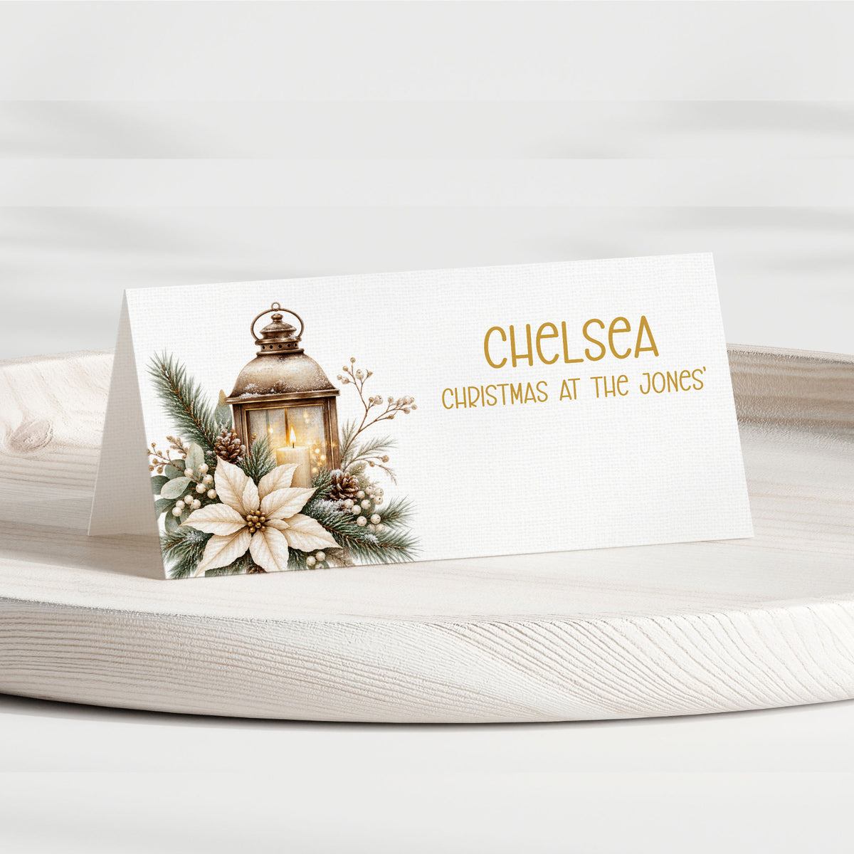 A personalised Christmas table place card on a white wooden tray. The card features a white background with a printed illustration of a lantern, pine branches, berries, and a white flower on the left, and the text 'CHELSEA CHRISTMAS AT THE JONES' in gold on the right.