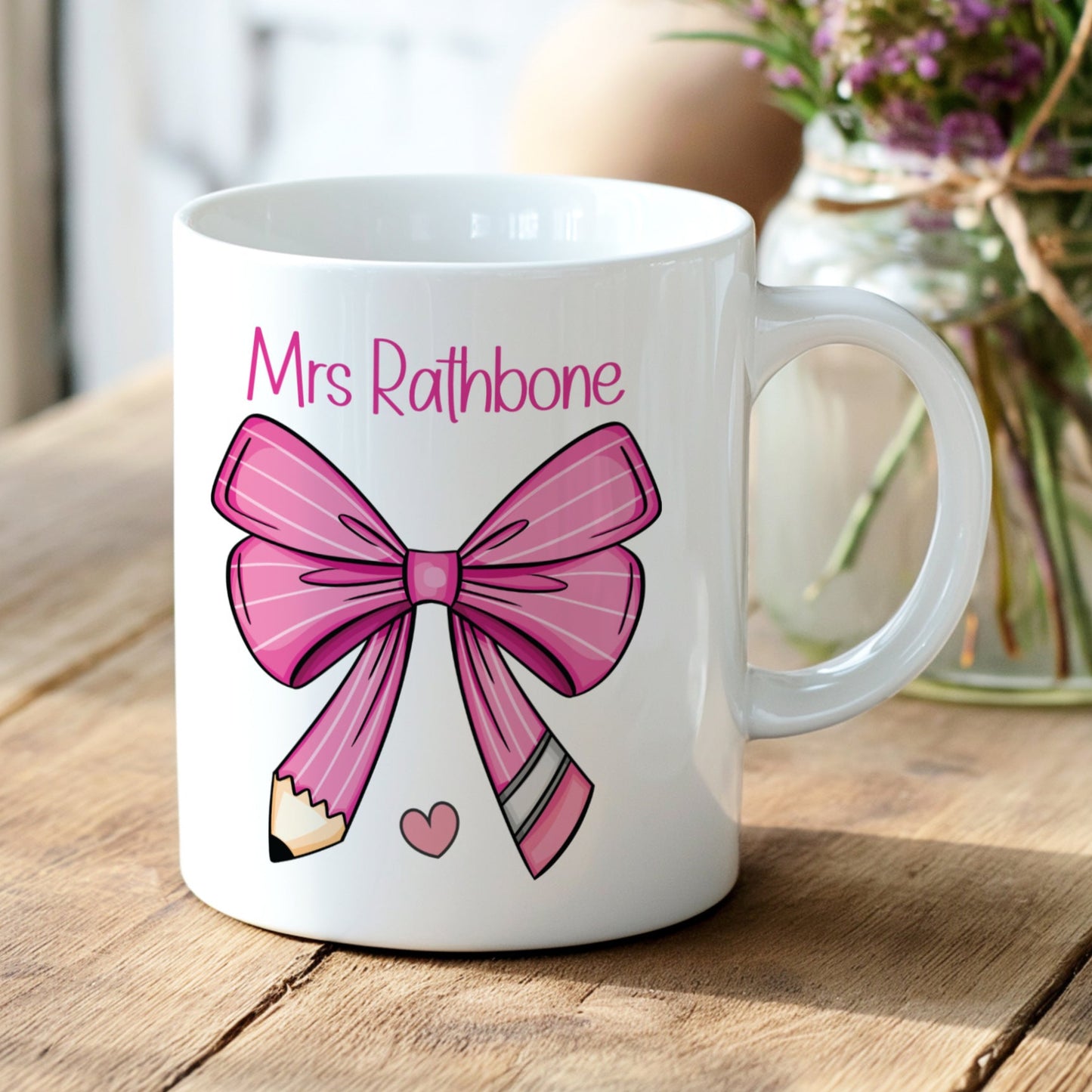 White mug with pink bow design and 'Mrs Rathbone' text on a wooden surface with flowers in the background