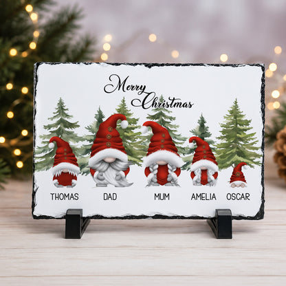 A rectangular slate Christmas slate on a black display stand, featuring illustrated Santa-clad gnomes labelled Thomas, Dad, Mum, Amelia, and Oscar with the text 'Merry Christmas' at the top. The sign is photographed on a light wood surface with blurred holiday lights and evergreen branches in the background.