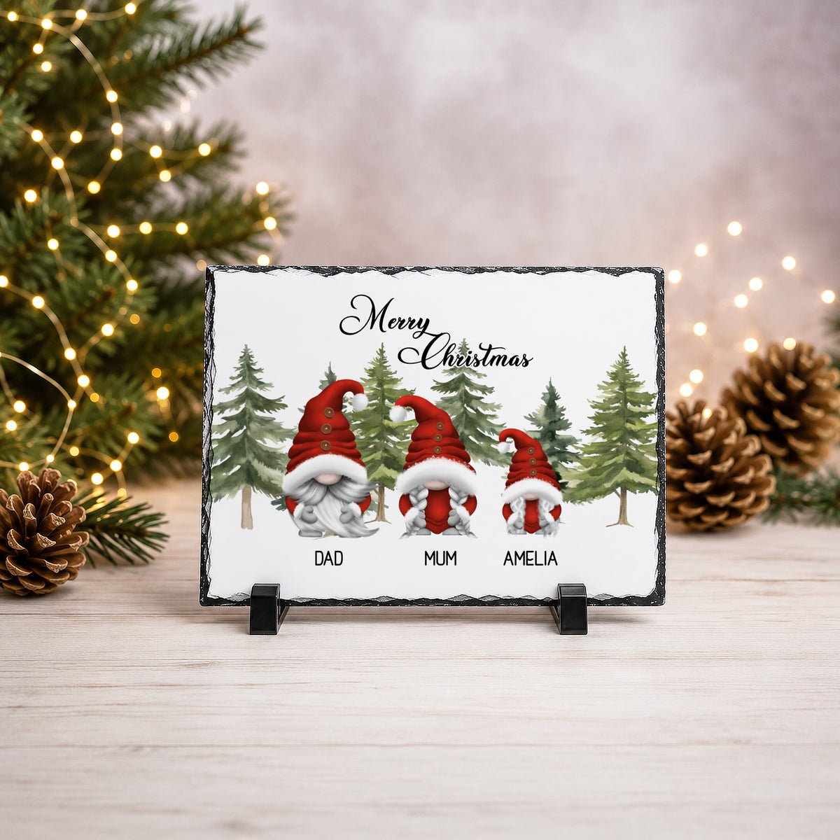 A rectangular slate Christmas sign on a small black stand, featuring three gnomes in red Santa hats and the text 'Merry Christmas' above them, with 'DAD', 'MUM', and 'AMELIA' printed below. The sign is photographed on a light wood surface with blurred Christmas trees, lights, and pinecones in the background.