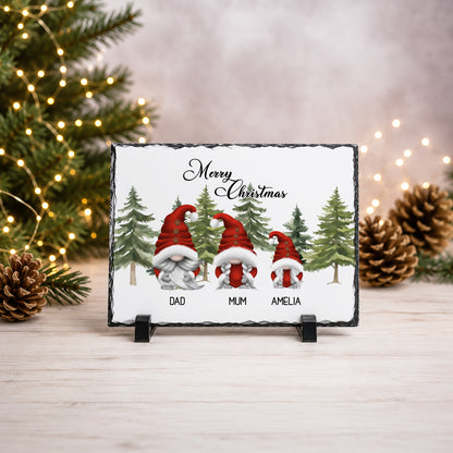 A rectangular slate Christmas sign on a small black stand, featuring three gnomes in red Santa hats and the text 'Merry Christmas' above them, with 'DAD', 'MUM', and 'AMELIA' printed below. The sign is photographed on a light wood surface with blurred Christmas trees, lights, and pinecones in the background.