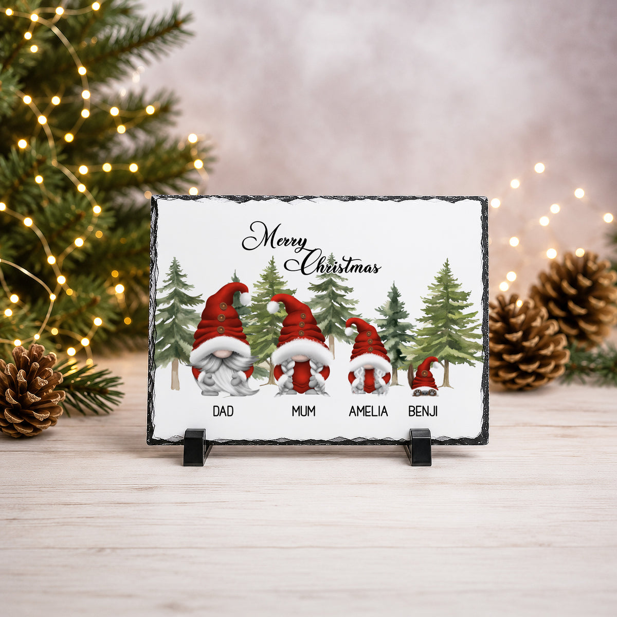 A rectangular slate Christmas plaque on a small black stand, featuring four gnomes in red Santa hats and coats with names printed below each figure. The plaque is photographed on a light wood surface with blurred Christmas tree branches, pinecones, and warm string lights in the background.
