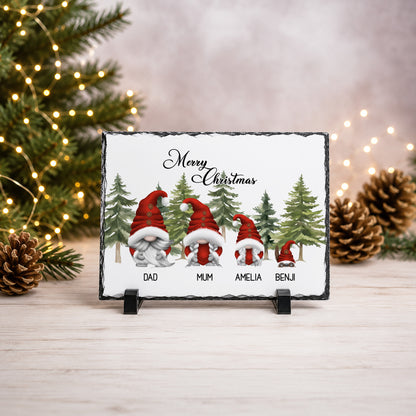 A rectangular slate Christmas plaque on a small black stand, featuring four gnomes in red Santa hats and coats with names printed below each figure. The plaque is photographed on a light wood surface with blurred Christmas tree branches, pinecones, and warm string lights in the background.