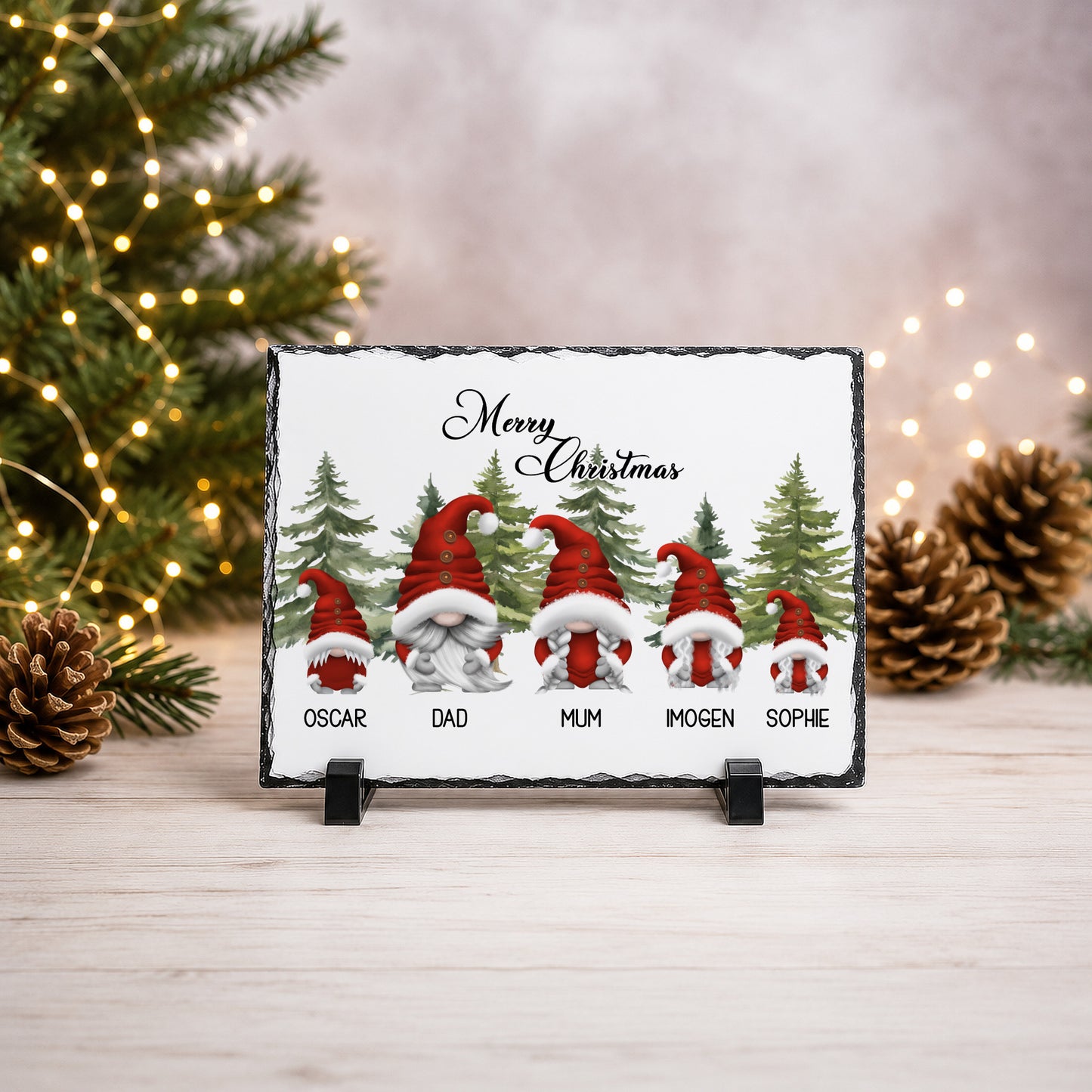 A rectangular slate Christmas sign on a small black stand, featuring five personalised gnomes with red hats and names underneath, with 'Merry Christmas' at the top. The sign is photographed on a light wood surface with blurred Christmas trees, pinecones, and warm string lights in the background.