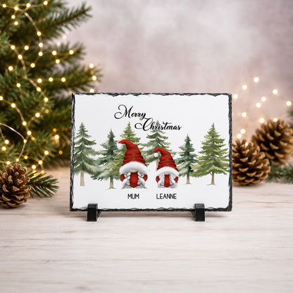 A rectangular slate Christmas plaque on a small black stand, featuring two gnomes in red Santa hats among green pine trees with the text 'Merry Christmas' and the names 'MUM' and 'LEANNE'. The plaque is styled on a light wood surface with blurred Christmas lights, pinecones, and a fir tree in the background.