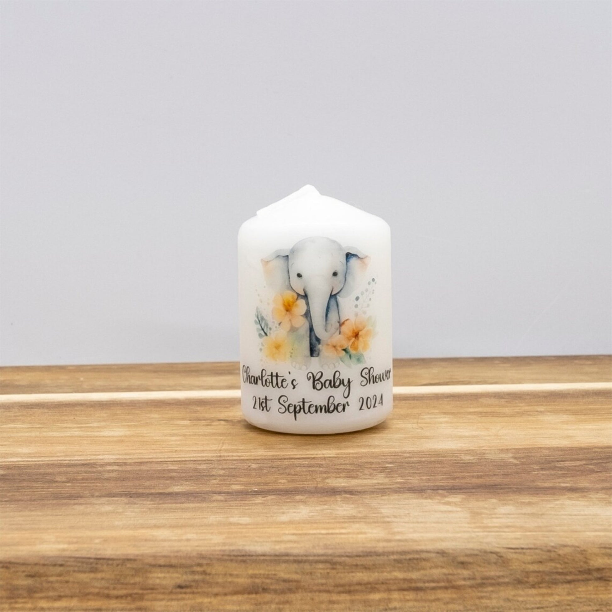 A personalised white pillar candle with a painted elephant and yellow flowers, sitting on a wooden surface against a neutral grey background.
