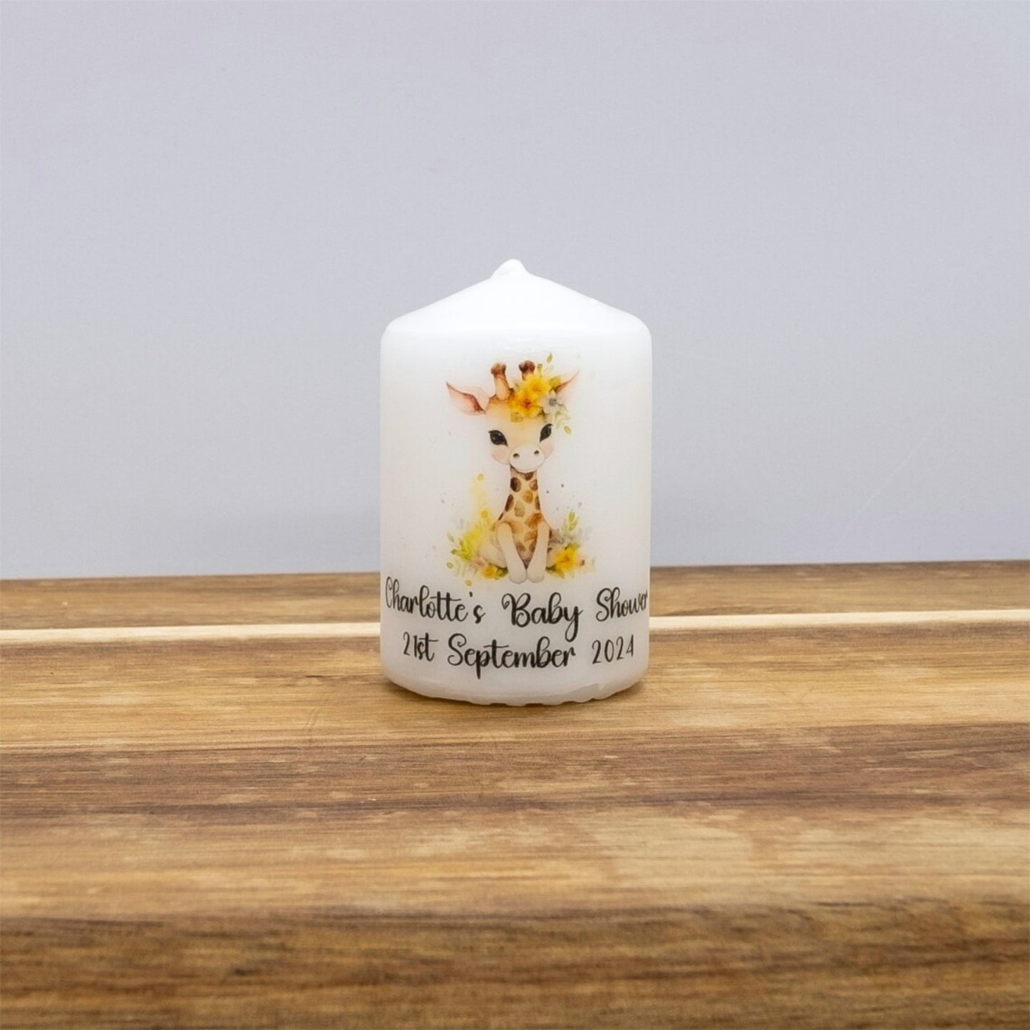 A personalised white taper candle featuring a watercolour giraffe illustration and the text 'Charlotte's Baby Shower 21st September 2024', displayed on a wooden surface against a neutral grey background.
