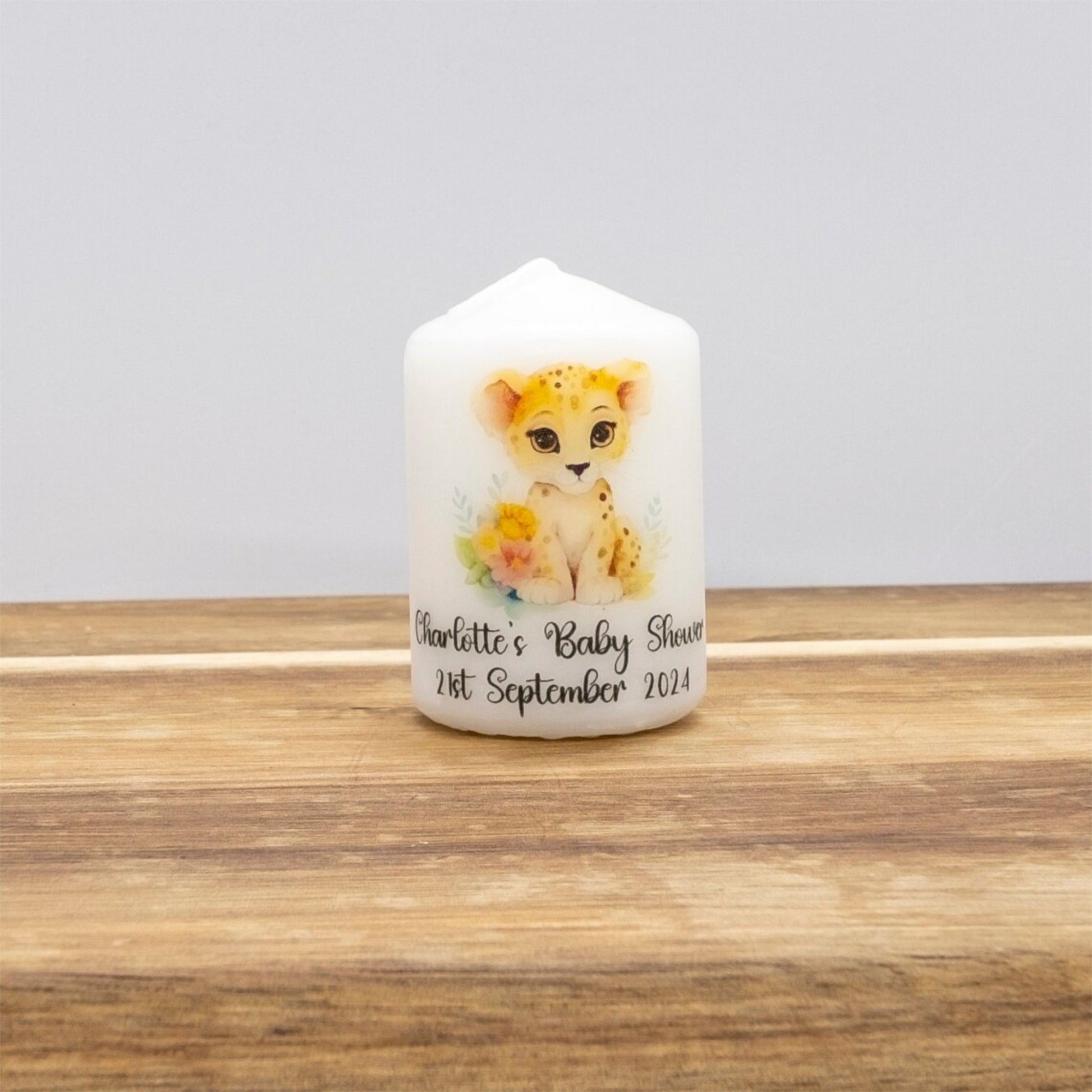 A white personalised house-shaped candle featuring a watercolour illustration of a yellow and black cheetah cub with flowers, and custom text reading 'Charlotte's Baby Shower 21st September 2024'. The candle is displayed on a wooden surface against a neutral grey background.