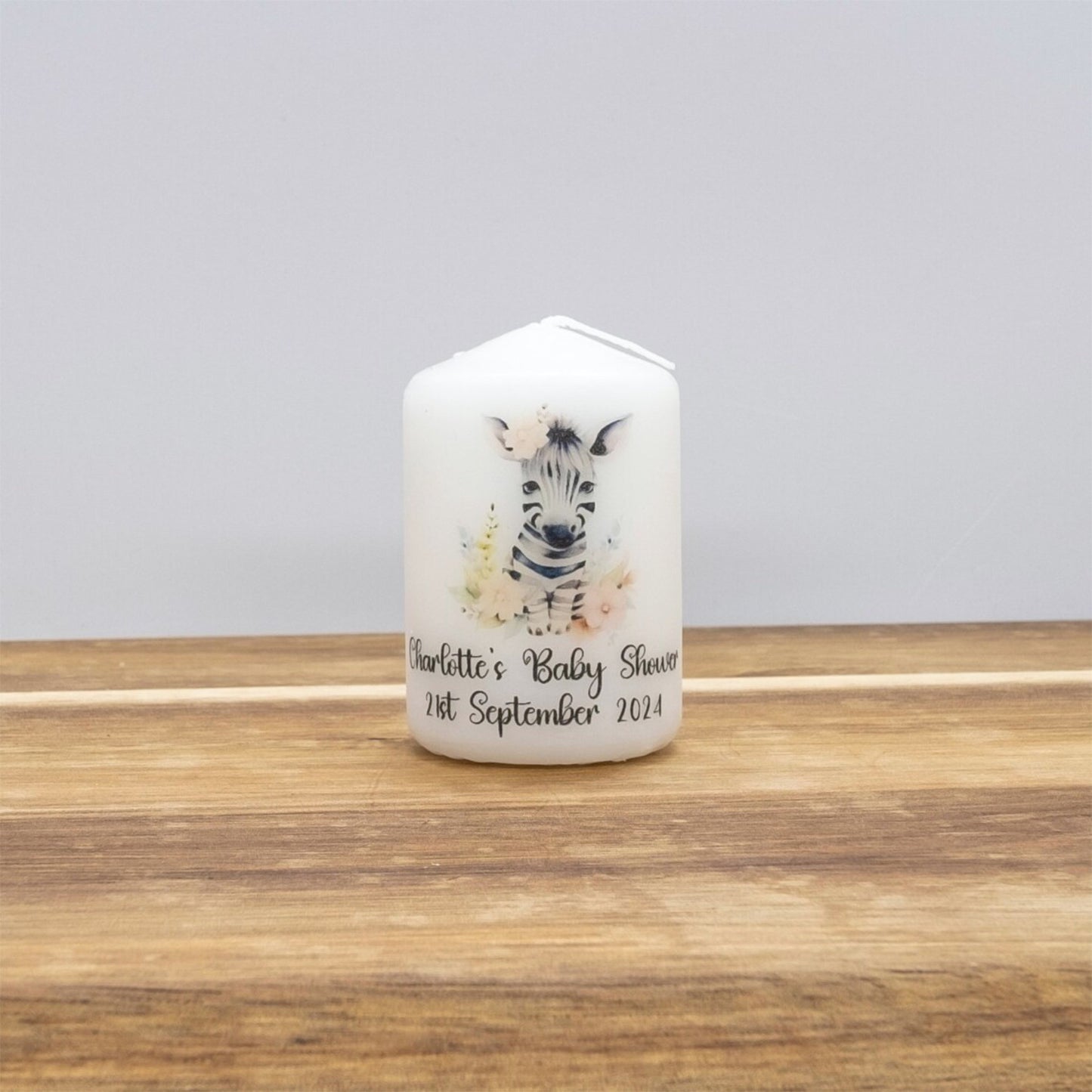 A personalised white pillar candle with a zebra illustration and custom baby shower text, standing on a wooden surface against a neutral grey background.