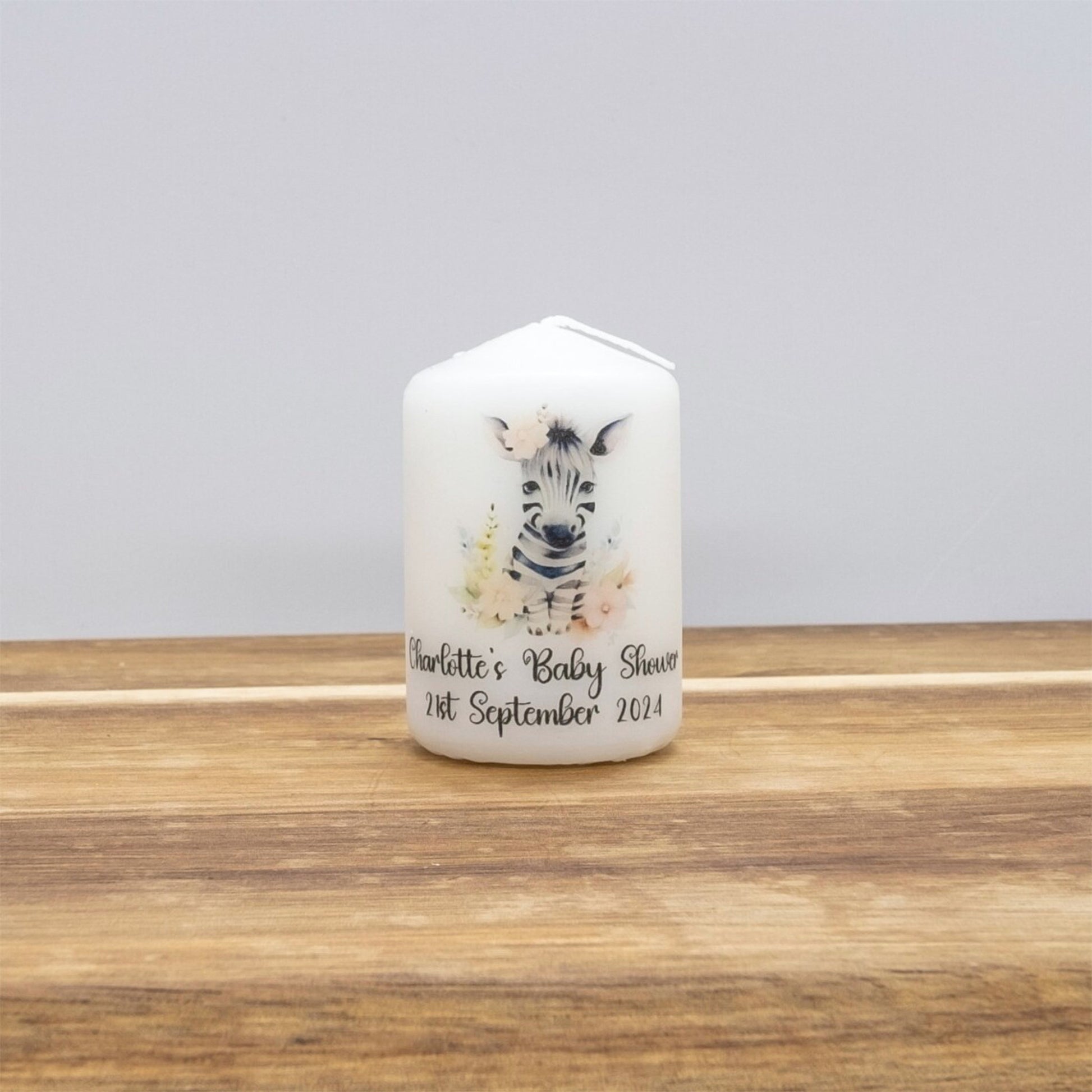 A personalised white pillar candle with a zebra illustration and custom baby shower text, standing on a wooden surface against a neutral grey background.