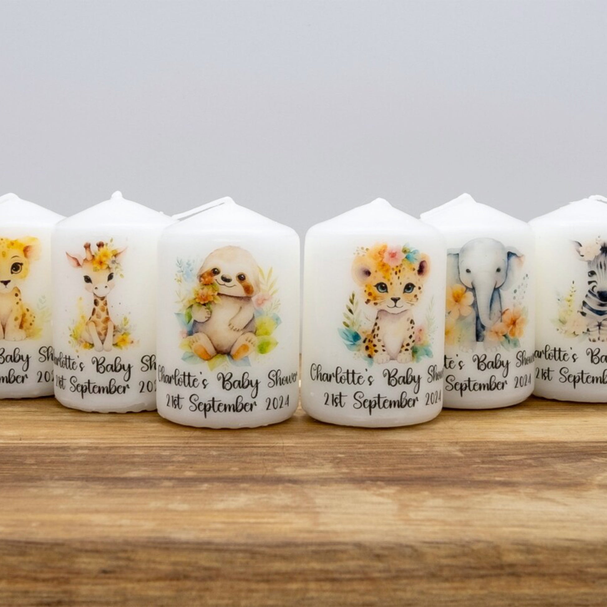 A row of personalised white taper candles featuring watercolour animal illustrations, including a giraffe, sloth, leopard, elephant, and zebra, with custom printed names and birth dates on a wooden surface against a light grey background.