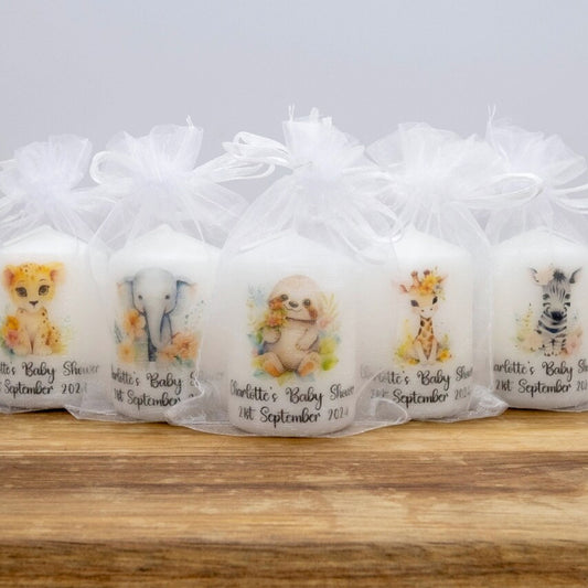 Five personalised baby shower favour candles in clear tulle bags, decorated with watercolour animal illustrations including a lion, elephant, dog, giraffe, and zebra, arranged on a wooden surface against a light grey background.