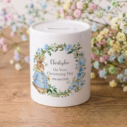 A white ceramic baby christening money box featuring a personalised illustration of Beatrix Potter's Peter Rabbit in a blue jacket, surrounded by a blue and green floral wreath. The box is printed with the name 'Christopher', 'On Your Christening Day', and a date.