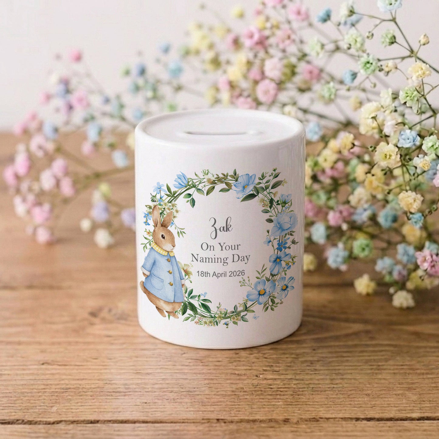A white ceramic piggy bank featuring a personalised illustration of Beatrix Potter's rabbit in a blue jacket, surrounded by a floral wreath and the text 'Jack On Your Naming Day 18th April 2025'. The piggy bank is set on a wooden surface with soft pastel baby's breath flowers in the background.