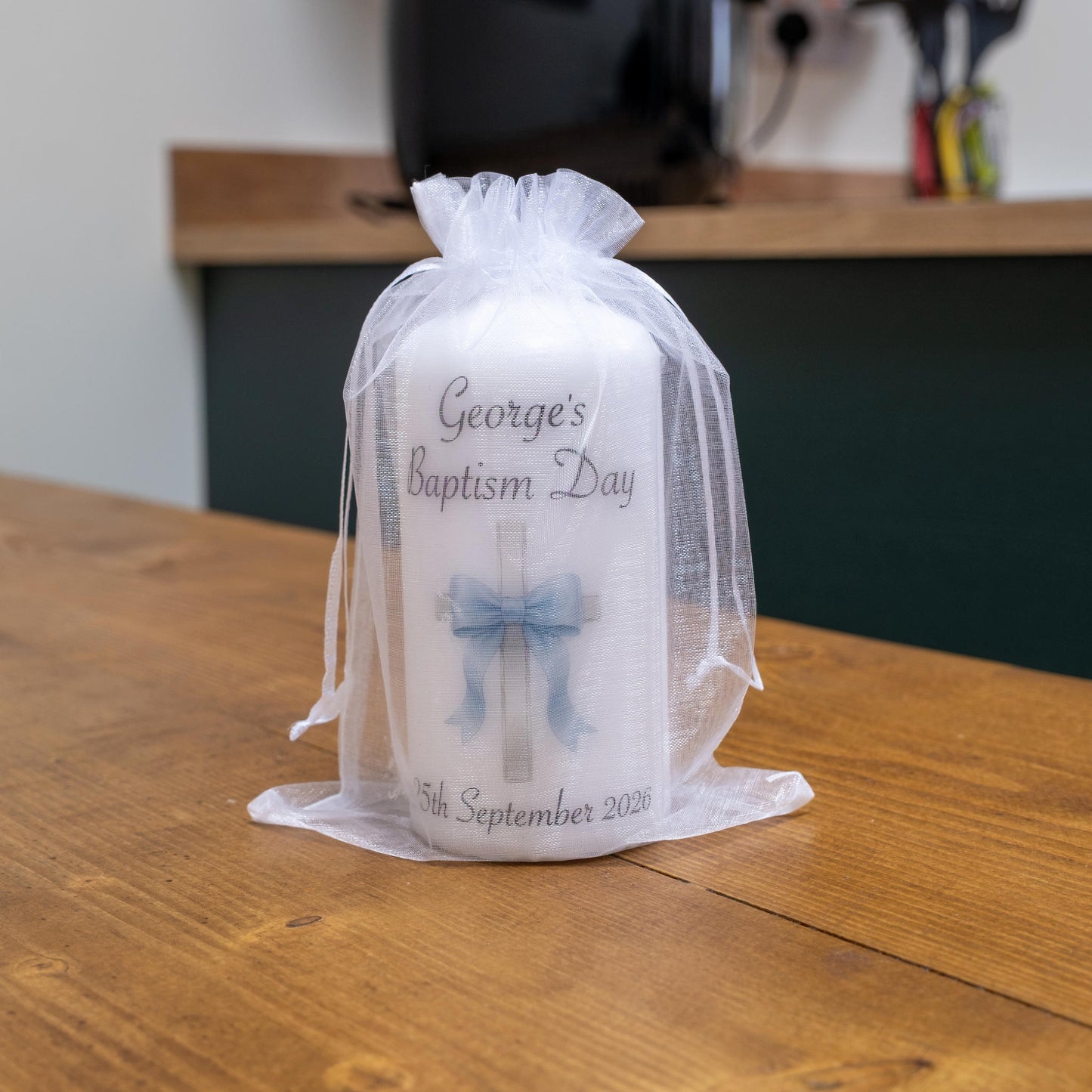 A white baptism candle wrapped in a sheer white organza bag, sitting on a wooden table. The candle features a printed cross with a blue bow and the text 'George's Baptism Day' and '5th September 2026'.
