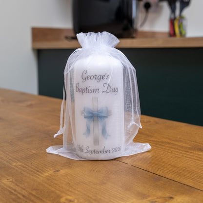 A white baptism candle wrapped in a sheer white organza bag, sitting on a wooden table. The candle features a printed cross with a blue bow and the text 'George's Baptism Day' and '5th September 2026'.