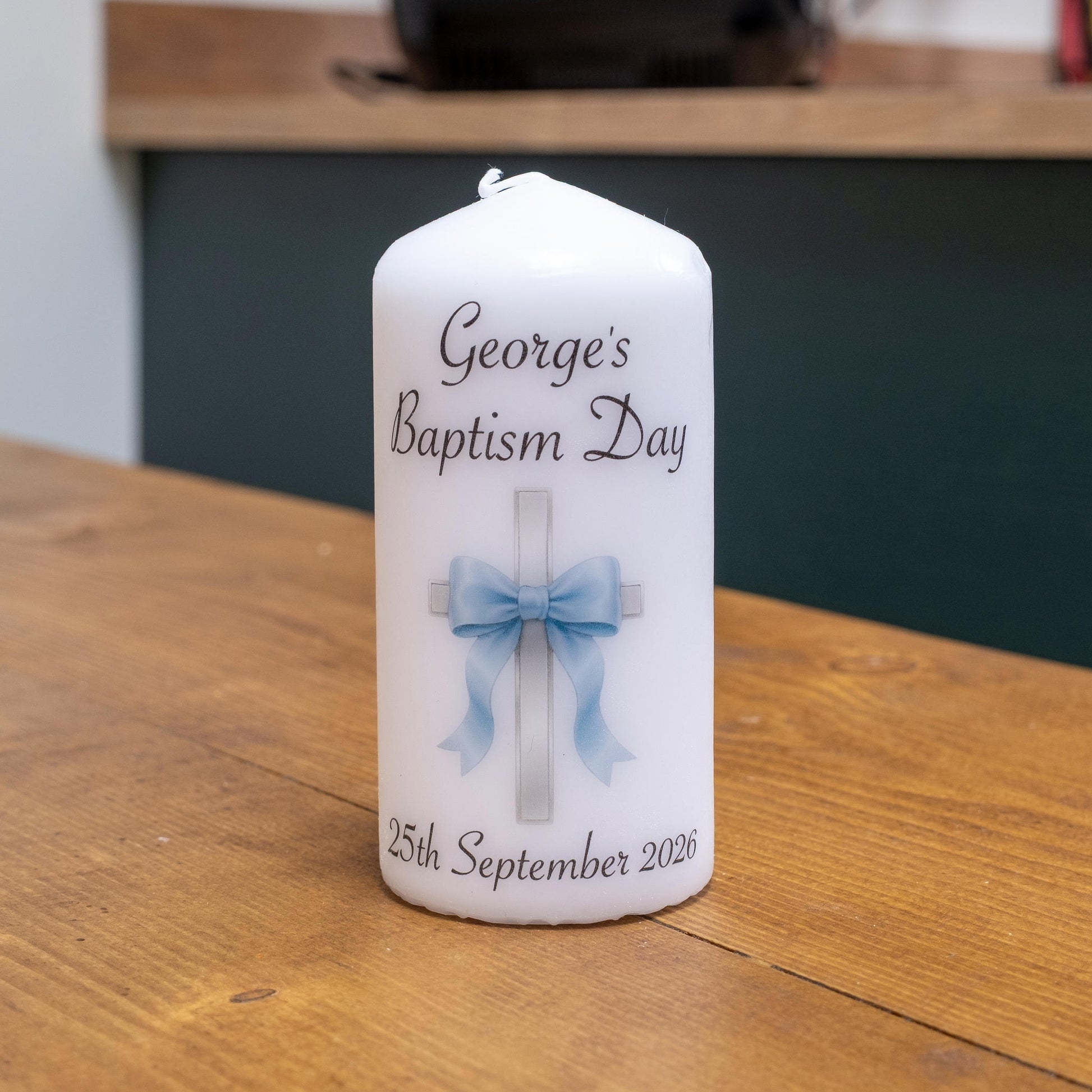 A white personalised birthday candle with 'George's Baptism Day' and '25th September 2026' printed on it, featuring a light blue bow and cross design, sitting on a wooden table.