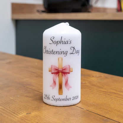 A personalised white pillar candle for Sophia's Christening Day, featuring a printed gold cross with a pink ribbon and the date 25th September 2026, set on a wooden table.