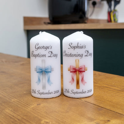 Two personalised white pillar candles on a wooden table. The left candle is labelled 'George's Baptism Day' with a blue ribbon and cross, and the right candle is labelled 'Sophia's Christening Day' with a pink ribbon and cross.