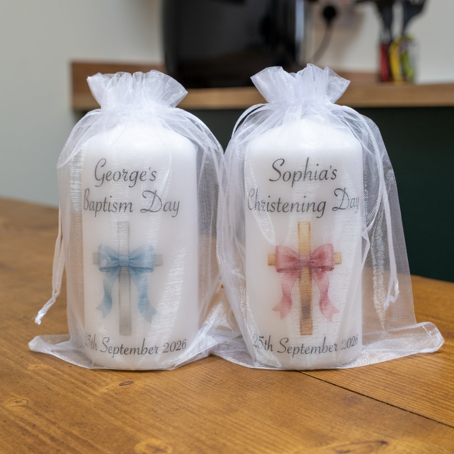 Two personalised white baptism candles wrapped in sheer white organza bags, displayed on a wooden table. The left candle is labelled 'George's Baptism Day' with a blue bow and cross graphic, and the right candle is labelled 'Sophia's Christening Day' with a pink bow and cross graphic.
