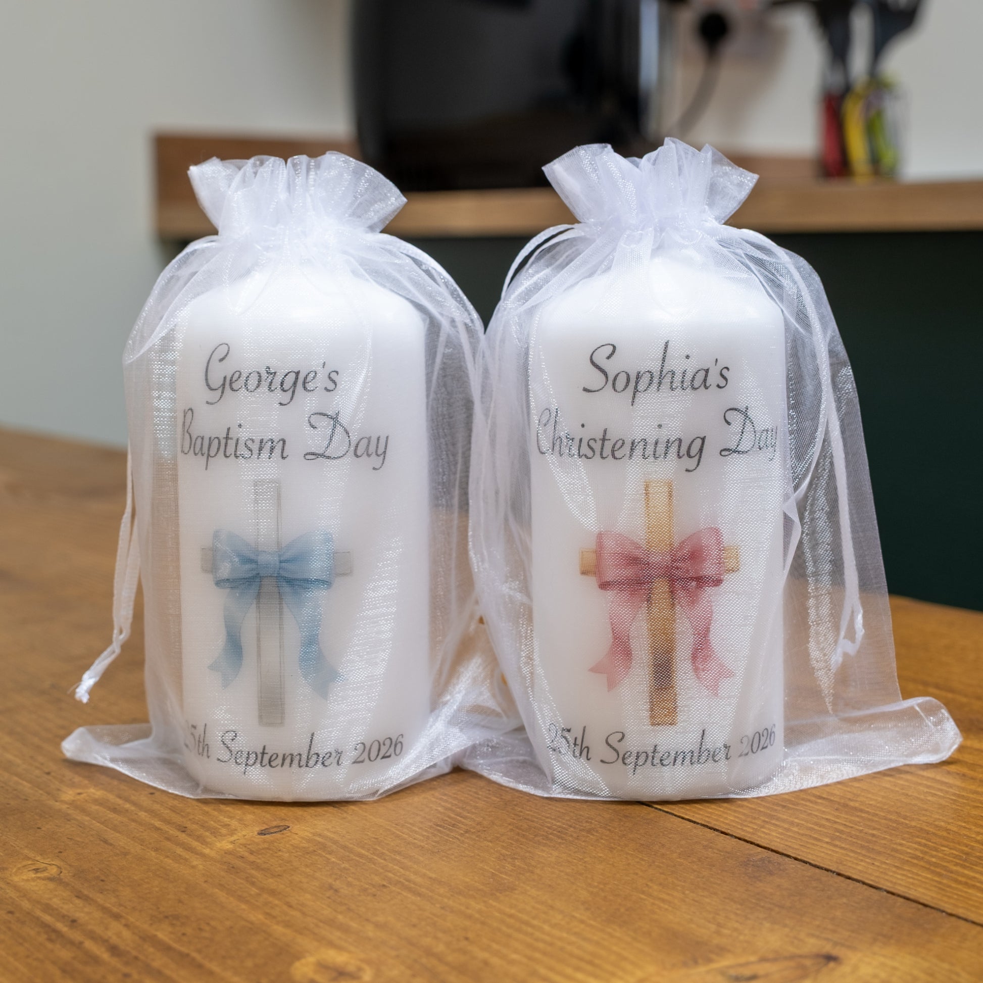 Two personalised white baptism candles wrapped in sheer white organza bags, displayed on a wooden table. The left candle is labelled 'George's Baptism Day' with a blue bow and cross graphic, and the right candle is labelled 'Sophia's Christening Day' with a pink bow and cross graphic.