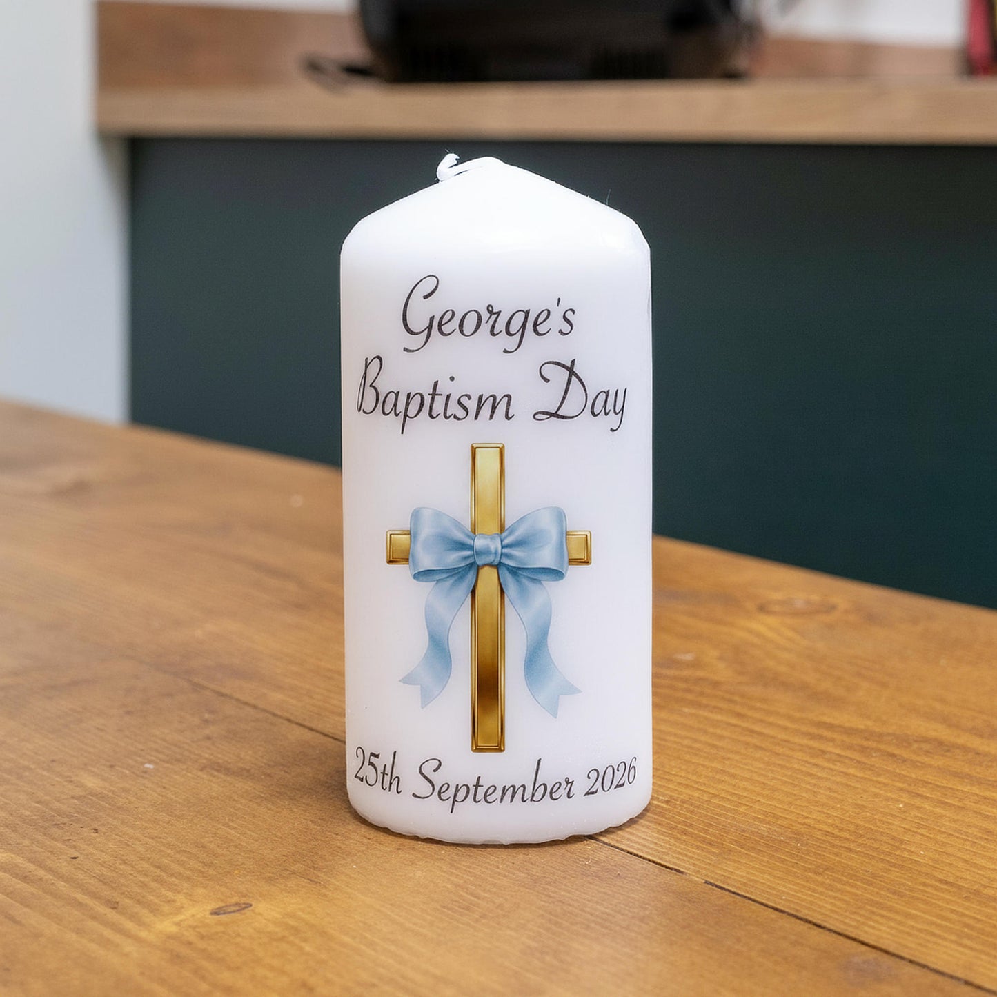 A white personalised baptism candle on a wooden table, featuring a gold cross with a light blue bow graphic and the text 'George's Baptism Day' and '25th September 2026'.