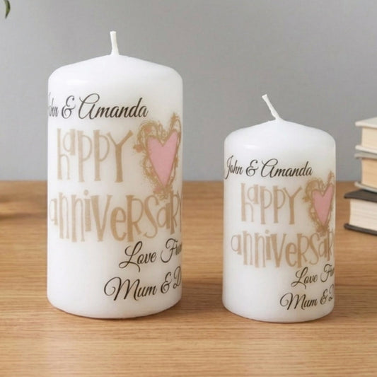 Two personalised white candles on a wooden table, one tall and one short, printed with 'John & Amanda', 'Happy Anniversary', and 'Love From Mum & Dad' in black and gold lettering with a pink heart design. Decorative flowers and books are in the background.