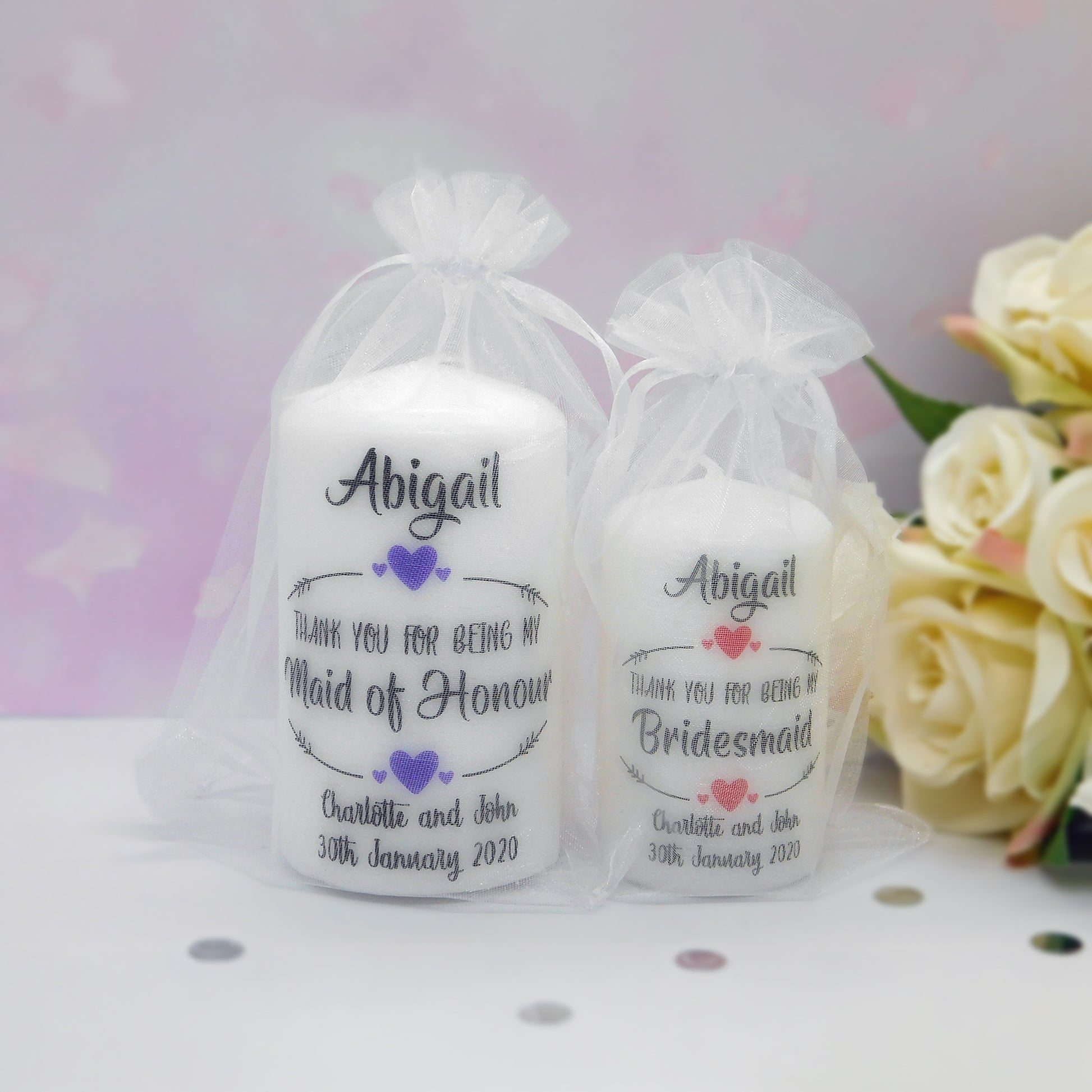 Two white taper candles in white organza gift bags with printed personalised text reading 'Abigail' and thank-you messages for Maid of Honour and Bridesmaid, shown on a light surface with cream roses in the background.
