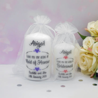 Two white taper candles in white organza gift bags with printed personalised text reading 'Abigail' and thank-you messages for Maid of Honour and Bridesmaid, shown on a light surface with cream roses in the background.