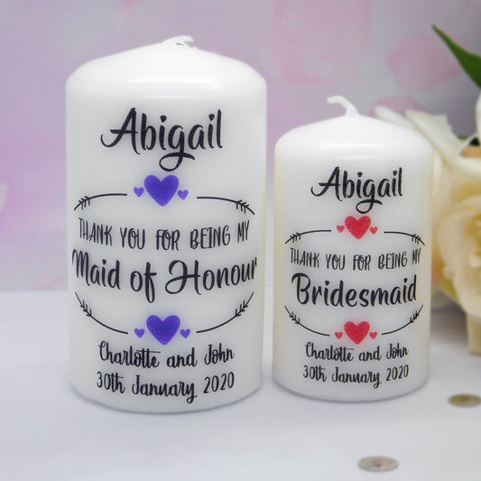 Two personalised white candles standing on a white surface, with a soft pink blurred background and white roses at the right. The larger candle reads 'Abigail' and 'Thank you for being my Maid of Honour' with purple hearts and names/date; the smaller candle reads 'Abigail' and 'Thank you for being my Bridesmaid' with red hearts and the same names/date.