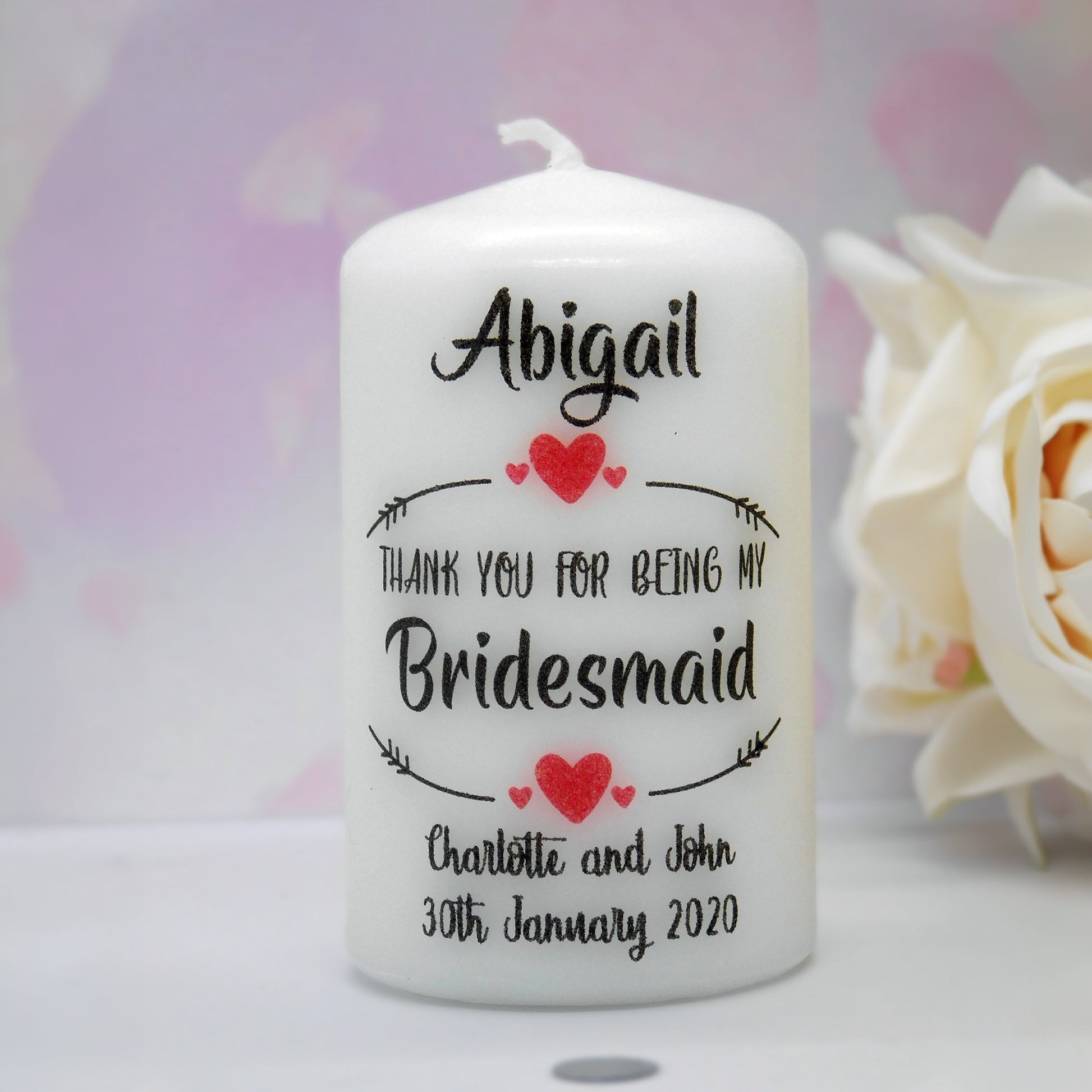 A personalised white pillar candle with black text reading 'Abigail Thank You for Being My Bridesmaid Charlotte and John 30th January 2020' and red heart accents, shown on a light surface with a blurred white flower and soft pink background.