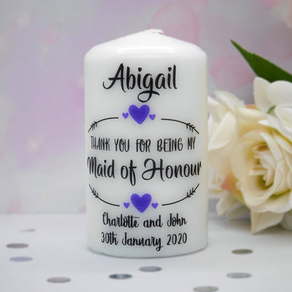 A white pillar candle with personalised black text and purple heart accents, reading 'Abigail Thank You for Being My Maid of Honour Charlotte and John 30th January 2020'. The candle is photographed on a white surface with blurred cream flowers in the background.