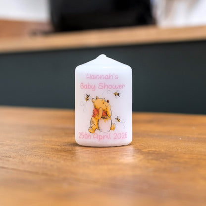 A custom white candle featuring Winnie the Pooh illustrations and the text 'Hannah's Baby Shower 25th April 2026' in pink, standing on a wooden table.