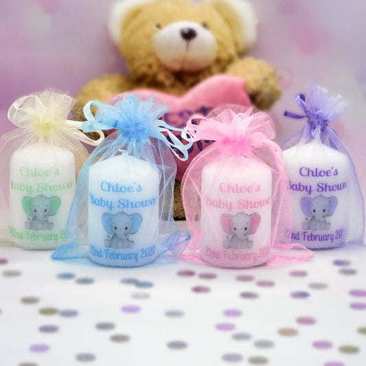 Four personalised baby shower favour bags in sheer tulle with bows, arranged in front of a teddy bear holding a pink heart. The bags are in white, light blue, pink, and lavender, each printed with a grey elephant illustration and the text 'Chloe's Baby Shower' and '2nd February 2020'.