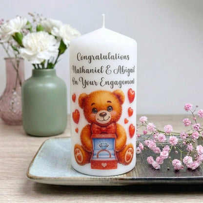 A white personalised candle with a brown teddy bear holding an engagement ring box and red hearts, with the text 'Congratulations Nathaniel & Abigail On Your Engagement!'. The candle is displayed on a tray with flowers in the background.