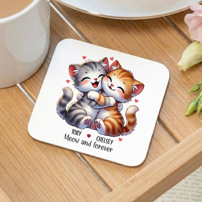 A square white coaster with rounded corners sits on a wooden table, printed with an illustration of two hugging cats and the text 'Toby + Chelsea Meow and forever' with small red hearts. A white mug, plate, and flowers are visible around it in a styled tabletop setting.