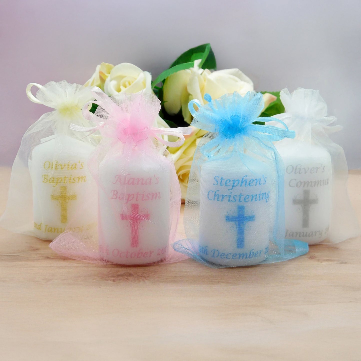 Four small white wax candles in sheer white organza bags tied with pastel ribbons, each printed with a cross and personalised baptism or communion text, arranged on a light wood surface with white roses in the background.