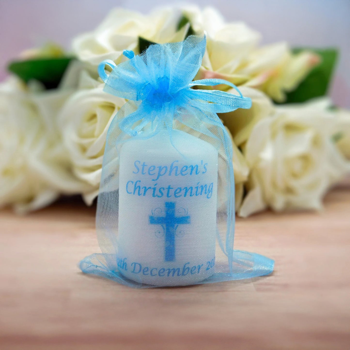 A personalised white candle wrapped in a sheer light blue organza drawstring bag, with blue text reading 'Stephen's Christening' and a blue cross, set against a blurred background of white roses.